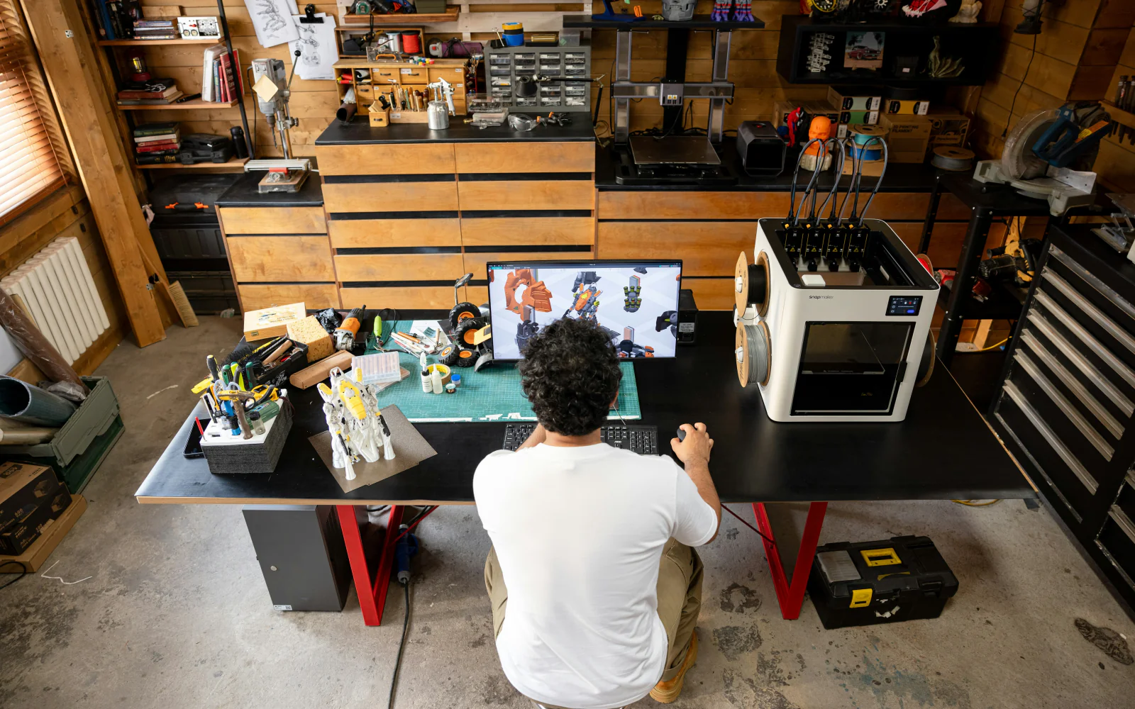 A designer working in CAD beside a 3D printer, viewed from above.